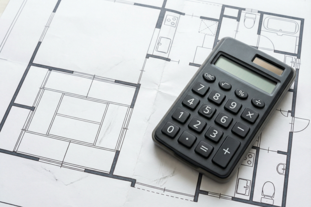 電卓と設計図の画像。本章では、最新の資材高騰や人件費上昇を反映した2026年のリノベーション費用を徹底解剖。中古住宅を刷新するフルリノベーションの平米単価や、キッチン・ユニットバス等の小規模な設備更新の相場を明確に提示する。築古物件の躯体補修や断熱・省エネ改修に伴う大規模な工事費用の目安、さらには住宅ローンや減税を賢く活用して実質負担を抑える方法まで網羅。資産価値を高め、理想の住まいを最適価格で手に入れるための最新情報をお届け